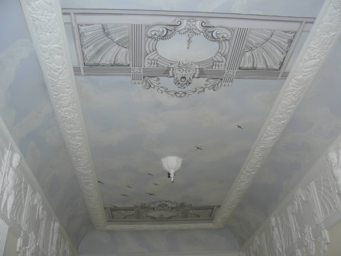 X Deckenmalerei als Eyecatcher in einem Entree aus dem Jugendstil. Das Ornament wurde findet sich im Stuck wieder und kann dem tektonischen Jugendstil zugeordnet werden. Der im Hintergrund befindliche Himmel gibt der Decke Tiefe und lässt die Graumalerei an der Decke plastisch wirken. Das Ornament selber wurde auch noch einmal an die Fassade angebracht. So findet es sich an 3 verschiedenen Bauteilen im Haus wieder. Zu dem eleganten aber prunkvollen Eingang wurde auch im Treppenhaus eine helle Marmormalerei / Marmorierung angebracht. An den Holzbauteilen findet sich eine Holzimitation in Mahagoni wieder. An der Fassade wurde auch eine Fassadenmalerei im Jugendstil angebracht. Die Firma restaurierung und gestaltung hat den Altbau gestaltet und alle Malerarbeiten mit seinem Team aus einem Restaurator, Maler und Malermeister umgesetzt.