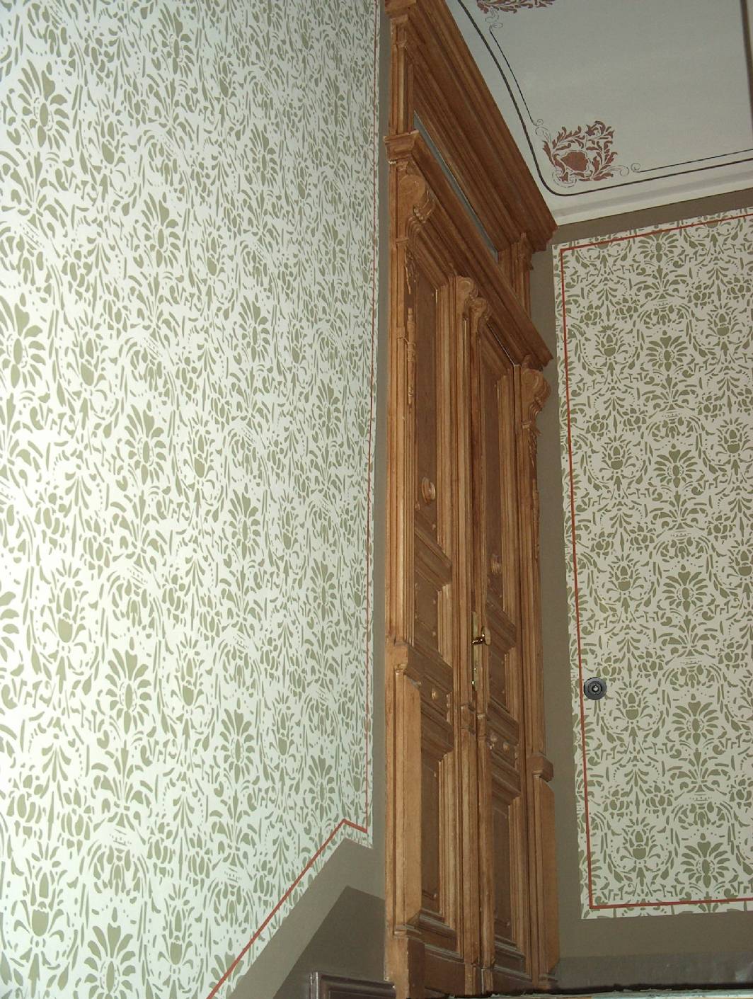 Zu sehen ist ein Ausschnitt von einem Altbau - Gründerzeit – Treppenhaus in Berlin. Als Deckenmalerei an den Podestdecken sind Begleitstriche und das Ornament als Schablonierung zu sehen. An den Wänden wurden Schablonen komplett angebracht und in Kassetten mit begleitstrich eigerahmt. An den Türflächen wurde eine Holzimitation / Holzmalerei in Form einer Bierlasur angearbeitet. Das Denkmal – Treppenhaus aus der Gründerzeit wurde komplett vom Eingangsbereich bis zum Treppenkopf rekonstruiert. Malermeister u. staatl. gepr. Techniker Norbert Schimpf arbeitet als Restaurator im Malerhandwerk und hat sich auf historische Malereien spezialisiert. Neben der Malerei übernimmt Malermeister Schimpf auch alle Vor-, Neben und Malerarbeiten im Altbau Treppenhaus. So kann das Unternehmen Treppenhausgestaltungen im Ganzen (z. Bsp. Sisal, Bakelit, Schaltern, Paneelleiste etc.) anbieten.