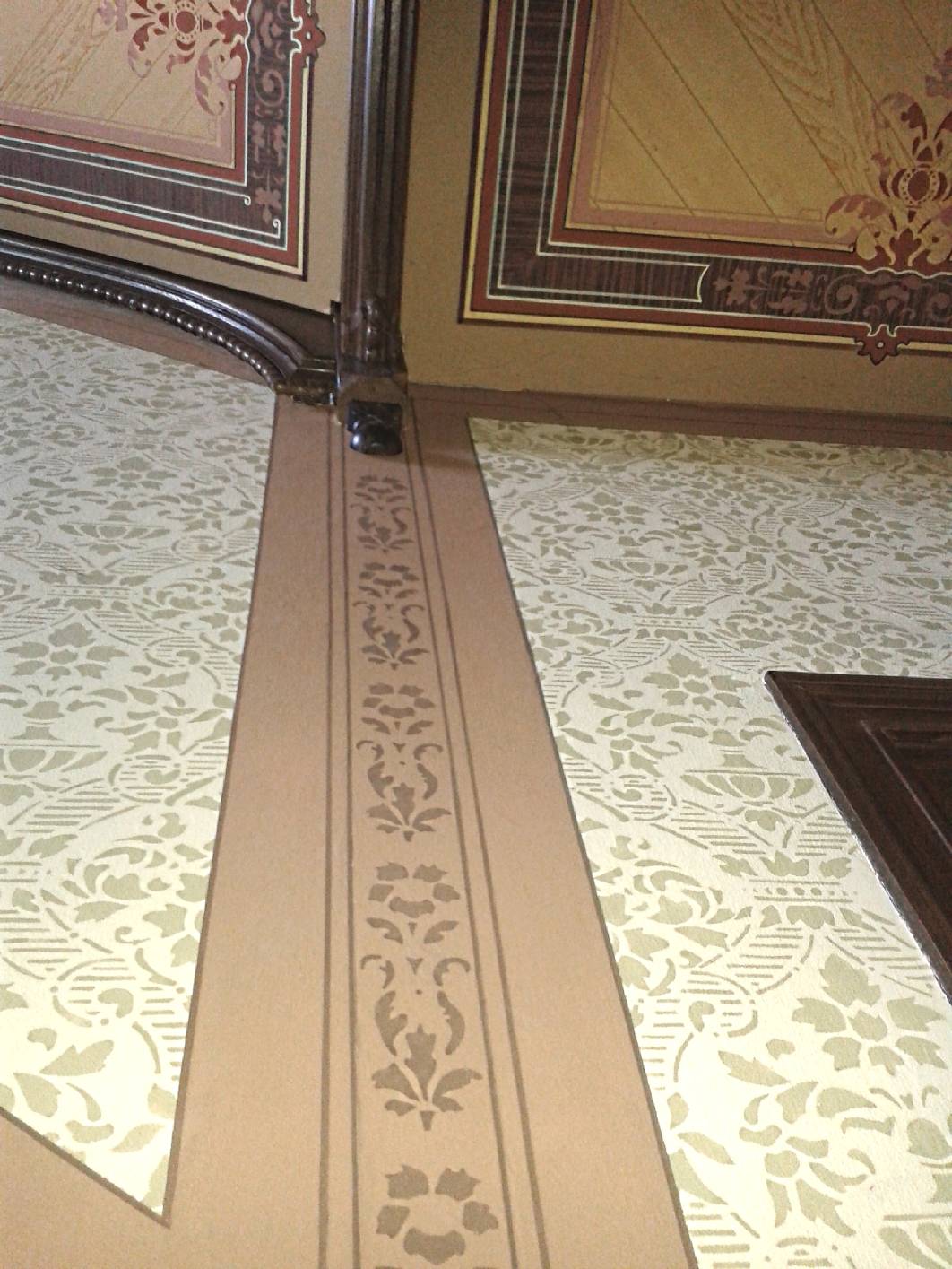 Ausschnitt eines Treppenhauses mit Deckenmalerei und Wandgestaltung in vollflächiger Schablonentechnik aus der Gründerzeit. An der Deckenmalerei ist eine gründerzeitliche Holzmalerei zu sehen. Die Wangen wurden wie die Türflächen mit einer Holzimitation in Lasurtechnik bemalt. Das Treppenhaus wurde vom Team Norbert Schimpf, der als Restaurator im Malerhandwerk das Projekt leitete, hergestellt. Vor der Gestaltung wurden die Malerarbeiten mit reversiblen Materialien angebracht. Jetzt leitet Norbert Schimpf seinen eigenen Betrieb. Er hat sich auf die Restaurierung im Altbautreppenhaus spezialisiert.